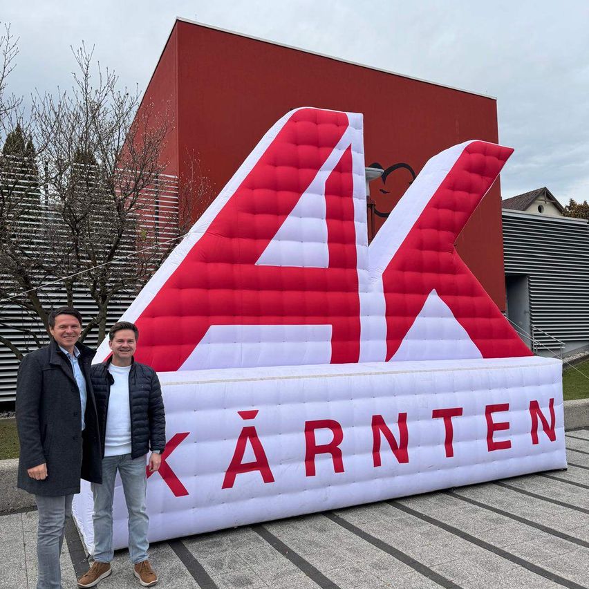 Two men stand next to an inflatable sign with the letters AK in red and white in front of a red building.