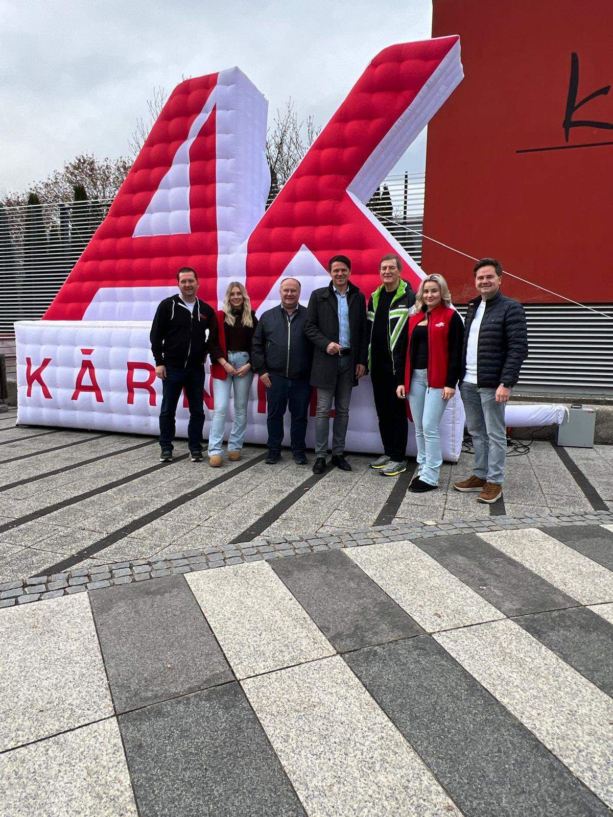 Six people stand in front of a large inflatable structure. The inflatable has a red and white design with the number 4 and the word Kärten in red. They are wearing winter clothing.