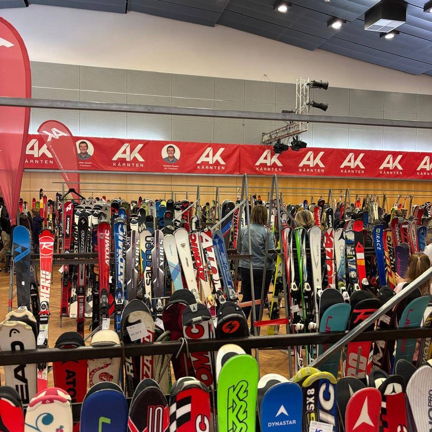 A large display of various skis is arranged in rows. A red banner with the letters AK and Karnten is displayed on the wall behind the skis. A person is standing in front of the skis.