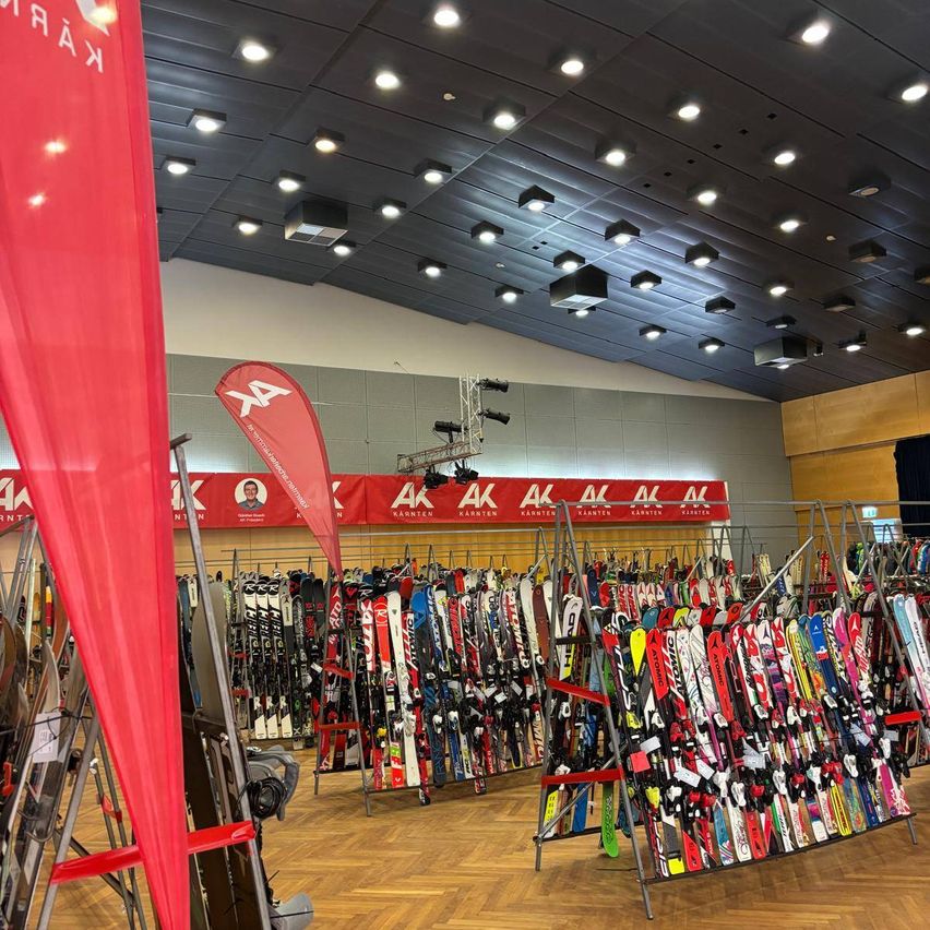 A ski shop with rows of skis and snowboards displayed. A red banner with white letters stands to the left. The ceiling has many recessed lights.