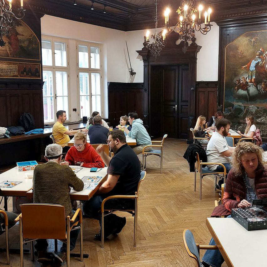 Eine Gruppe von Menschen spielt Brettspiele in einem Raum mit Holzböden und Kronleuchtern. Einige sitzen, andere stehen, mit verschiedenen Spielbrettern sichtbar.