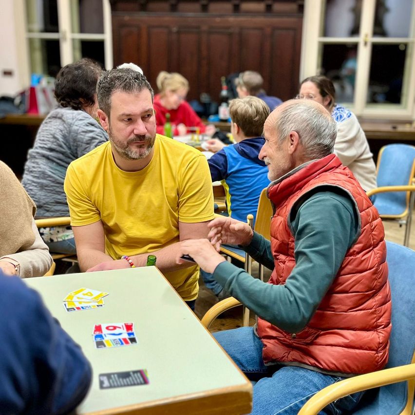Zwei Männer sitzen an einem Tisch und spielen ein Brettspiel. Der Mann in Gelb gestikuliert mit der Hand. Der ältere Mann in Rot konzentriert sich auf das Spielbrett.
