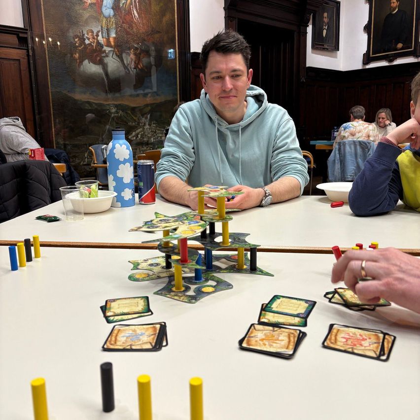 Ein Mann sitzt an einem Tisch mit einem Brettspiel. Verschiedene farbige Marker und Karten sind auf dem Tisch. Er sieht ernst aus, während er spielt.