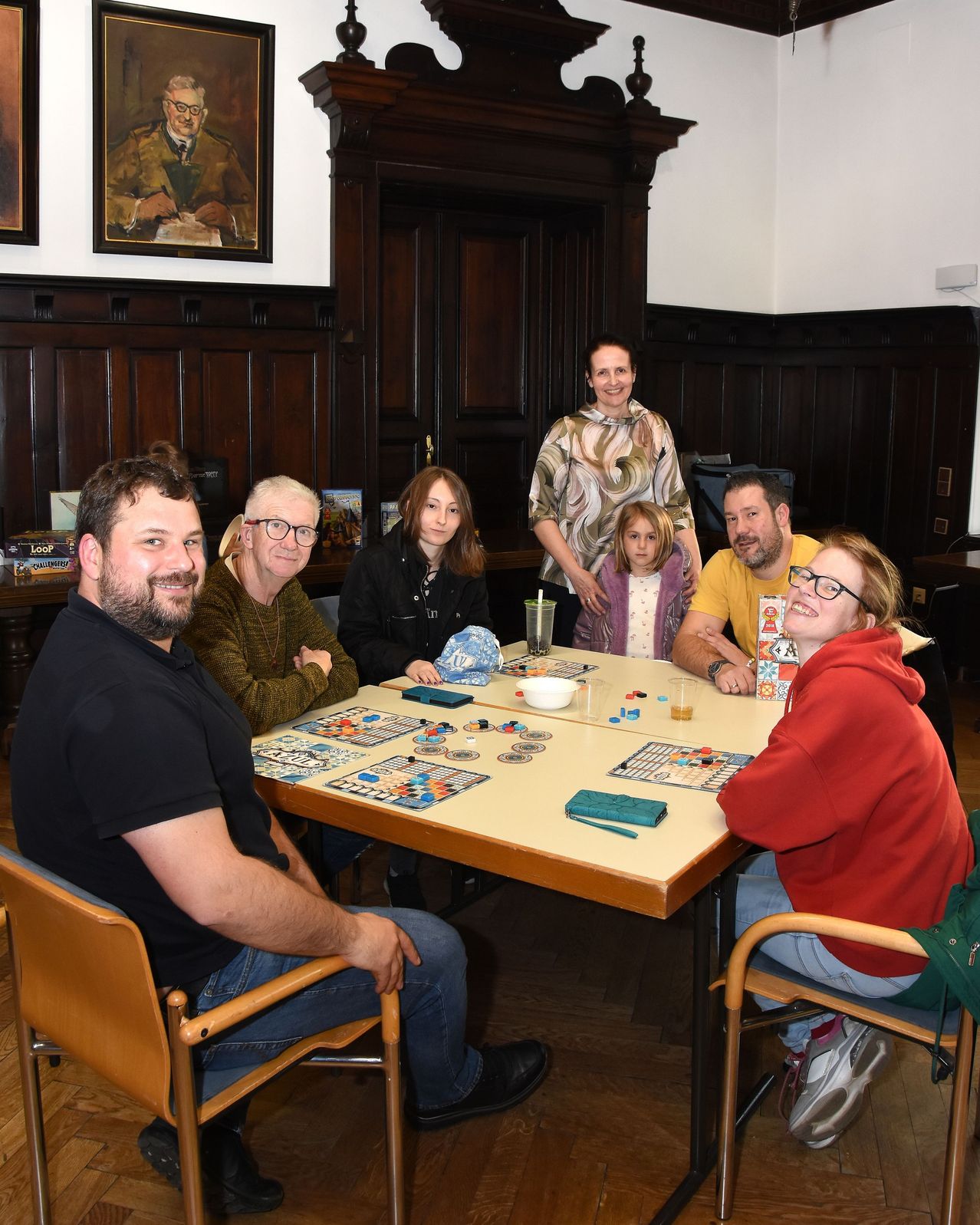 Eine Gruppe von Menschen sitzt um einen Tisch und lächelt. Sie spielen ein Brettspiel mit einem Glas Wasser und einem Becher auf dem Tisch.