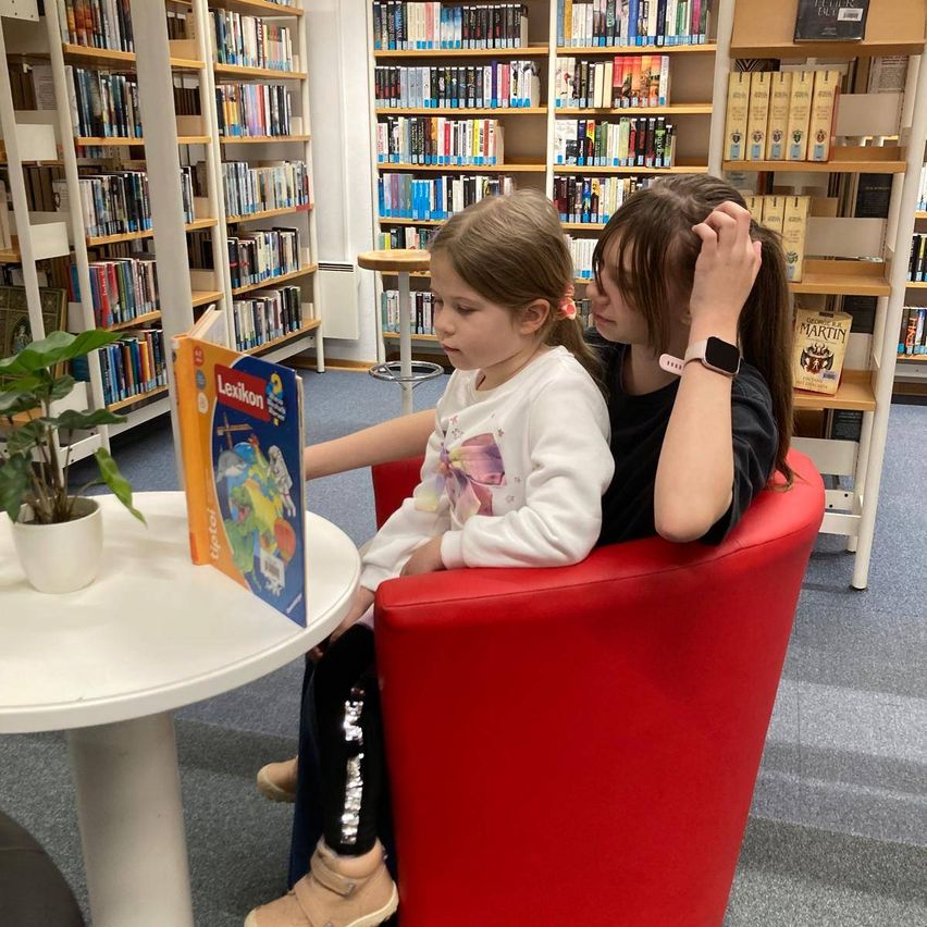 Zwei junge Mädchen sitzen in einer Bibliothek, eine liest ein Buch, während die andere ihren Kopf hält. Vor ihnen steht ein kleiner Tisch mit einer Pflanze.