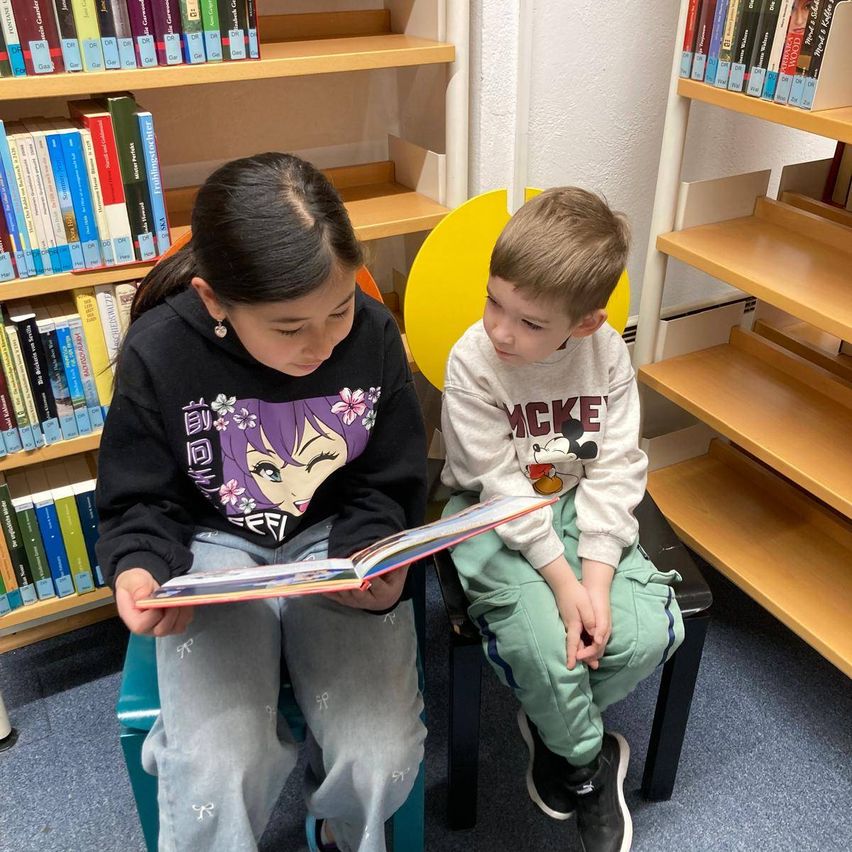 Ein Mädchen und ein Junge sitzen auf Stühlen in einer Bibliothek. Das Mädchen liest dem Jungen ein Buch vor.