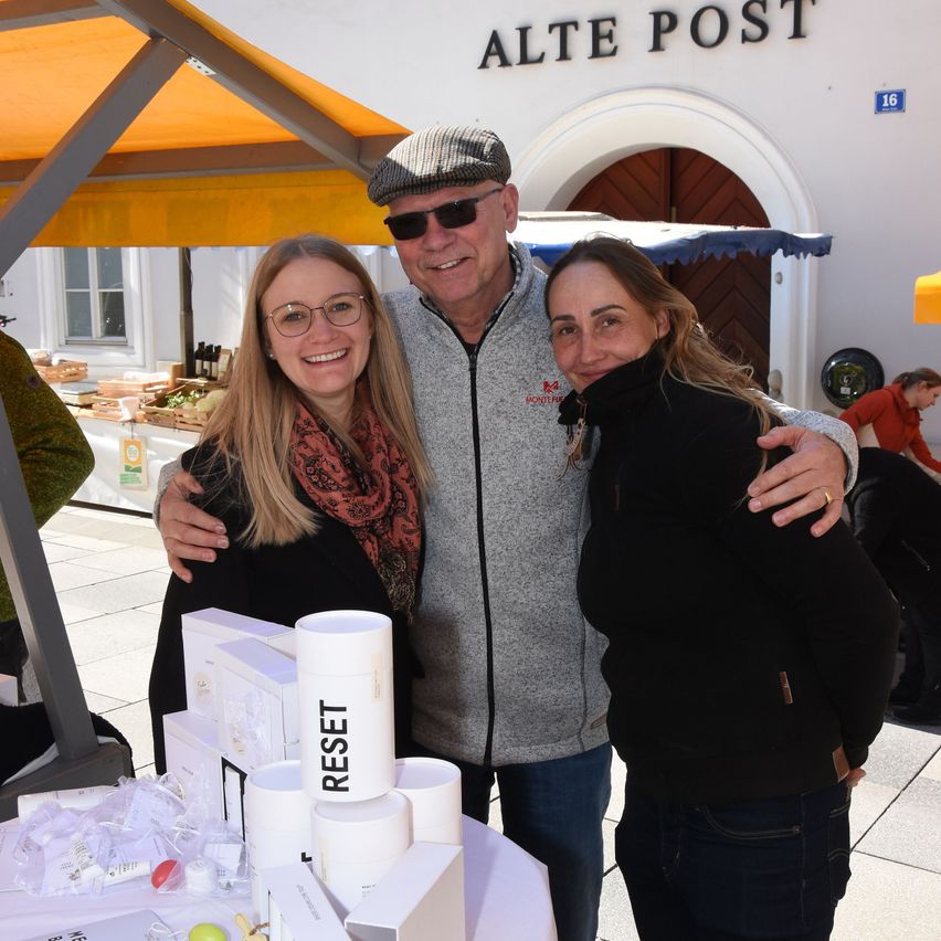 Drei Personen stehen an einem Tisch mit Produkten. Der Mann trägt einen Hut und Sonnenbrille. Die Frauen tragen Brillen und Schals. Der Tisch hat mehrere Kisten und ein Produkt mit der Aufschrift 'RESET'. Hinter ihnen steht ein Gebäude mit dem Namen 'Alte Post'.