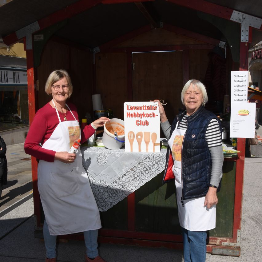 Zwei Frauen in Schürzen stehen neben einem Essensstand. Sie lächeln und halten ein Schild mit der Aufschrift 'Lavantaler Hobbykoch-Club'.