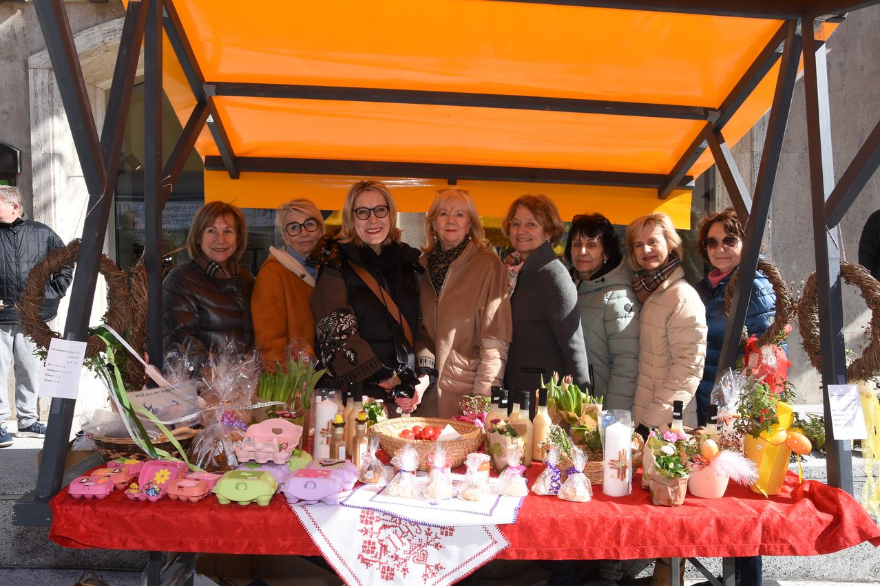 Eine Gruppe von Frauen steht hinter einem Tisch voller Osterdekorationen. Jede Frau trägt Winterkleidung und sie alle scheinen zu lächeln.