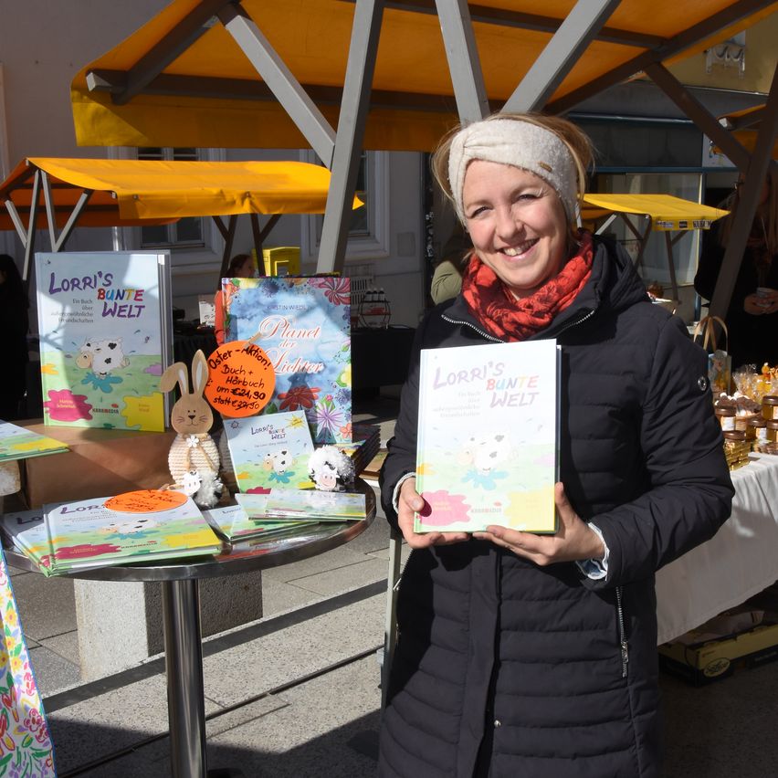 Eine lächelnde Frau steht im Freien und hält ein Buch mit dem Titel Lorris Bunte Welt, umgeben von Büchern und Spielzeug auf einem Tisch.