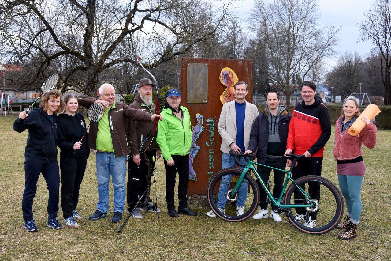 Sieben Menschen stehen im Freien, zwei halten Sportgeräte, einer ein Fahrrad, und eine Wand mit Graffiti im Hintergrund.