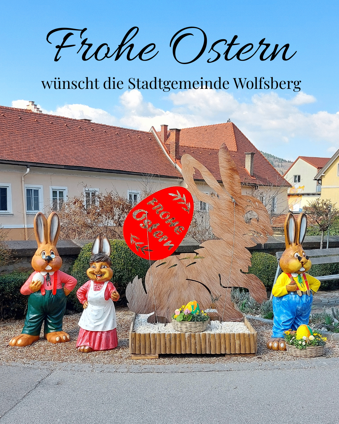 Three bunny statues stand outside a house with a red sign that says 'Frohe Ostern' and 'Wolfsberg Gemeinde' on top. A large rabbit statue is in front of them.