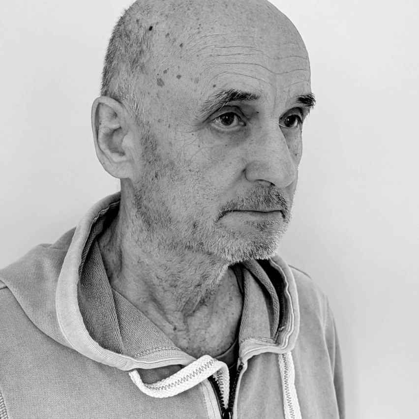 An older man with a bald head, a beard, and a mustache is wearing a gray hoodie jacket with a zipper. He looks serious and focused.