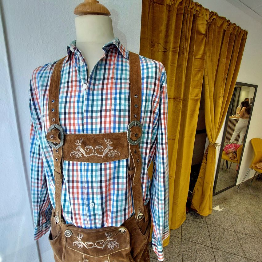 A mannequin displays a colorful checkered shirt with brown leather suspenders with silver buckles. It is placed against a white wall with a curtain in the background.