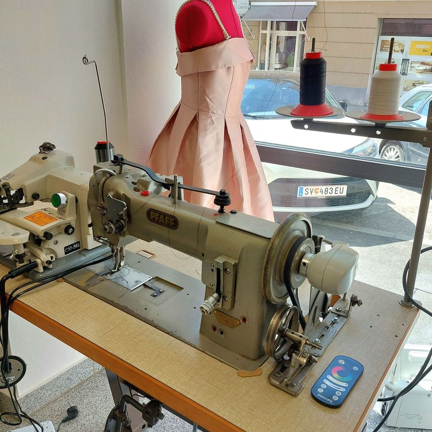 An industrial sewing machine on a wooden table, with a remote control, a pink dress mannequin in the background, and spools of thread. A white car is parked outside.