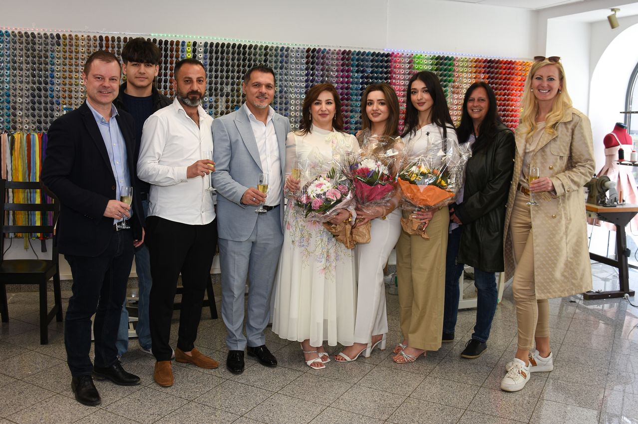A group of people pose for a photo in front of a colorful wall with flowers in hand. Some hold wine glasses.