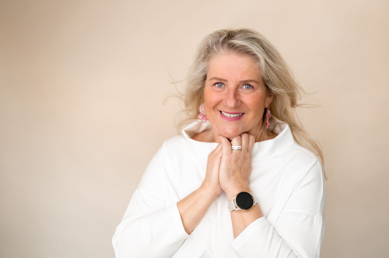 A woman with gray hair wearing a white shirt and earrings smiles, holding her hands on her chest. She wears a silver ring and a black smartwatch.