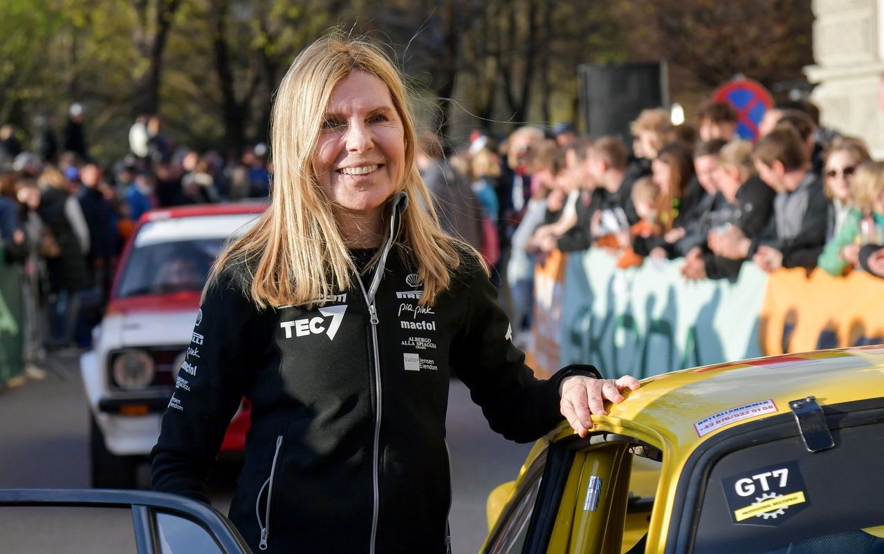 A woman with blonde hair wearing a black jacket with white text and logos stands beside a yellow car, smiling and posing for a photo. Behind her, a crowd of people watches, some with blurred faces.
