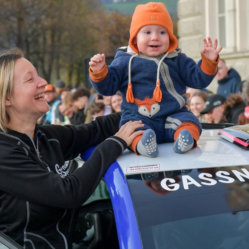 A woman holds a baby on top of a blue car. The baby is dressed in blue with a fox design. The car has the word Gassner on it.