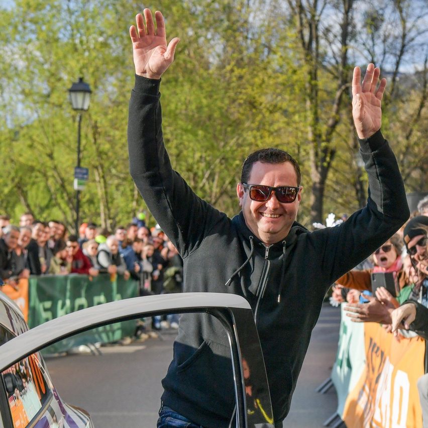 A man waves to a crowd while standing in front of a car with a banner behind him. He wears a black hoodie and sunglasses.