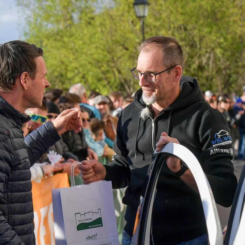 Two men are shaking hands. One holds a white bag. The other wears a black hoodie with 'Welvic' and 'Allianz' on it. They are in front of a crowd.