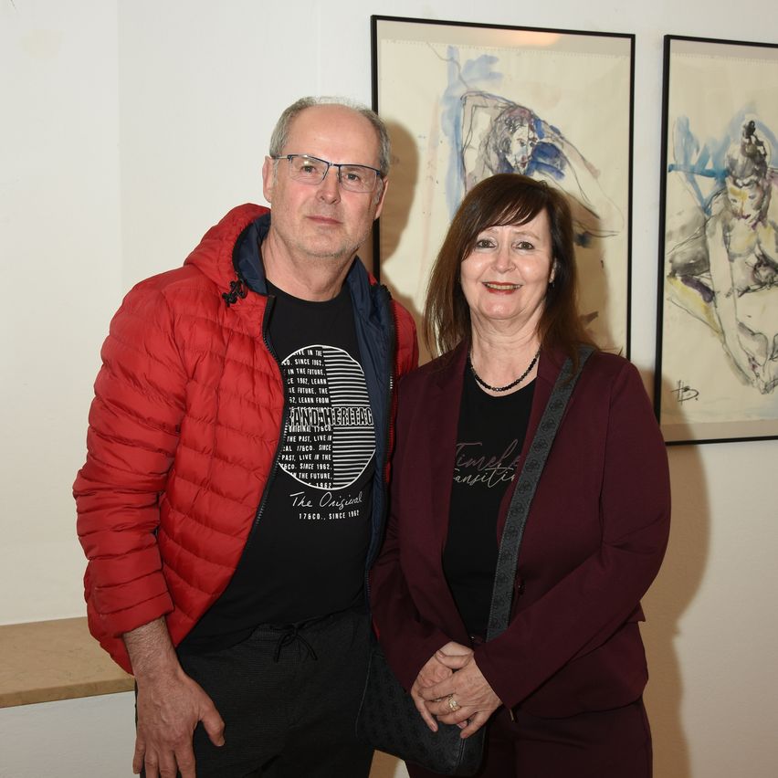 An older man and woman stand next to each other in front of an art exhibit. The man wears a red jacket, and the woman wears a maroon jacket. Both are smiling.
