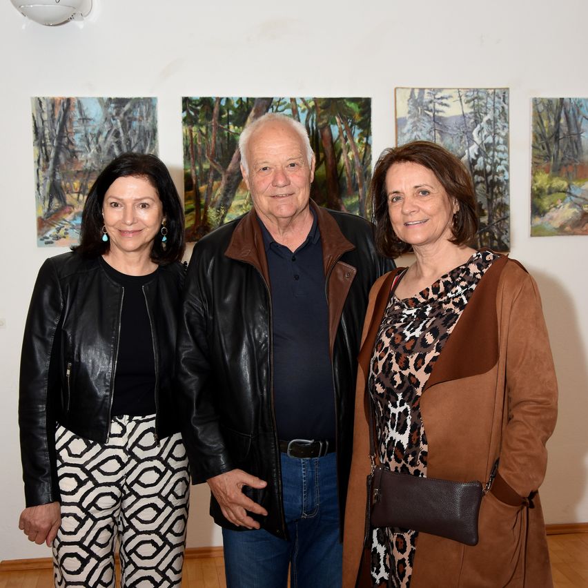 Three people stand close together in an art gallery. They are smiling and appear to be posing for a photograph. The walls are adorned with several paintings.