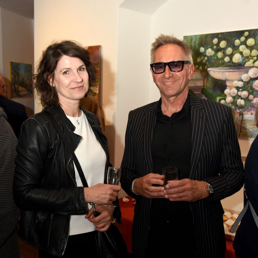 An adult man and woman are standing next to each other in an art gallery, holding glasses. Behind them, various paintings are on the walls, and other people are standing in the background.