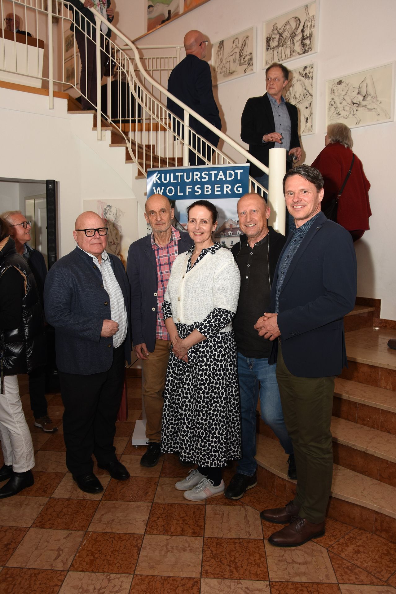 A group of people stands on a staircase, smiling and looking at the camera. Behind them, a sign reads 'Kulturstadt Wolfsberg.' The setting is indoors, with a staircase, walls, and a door in the background.