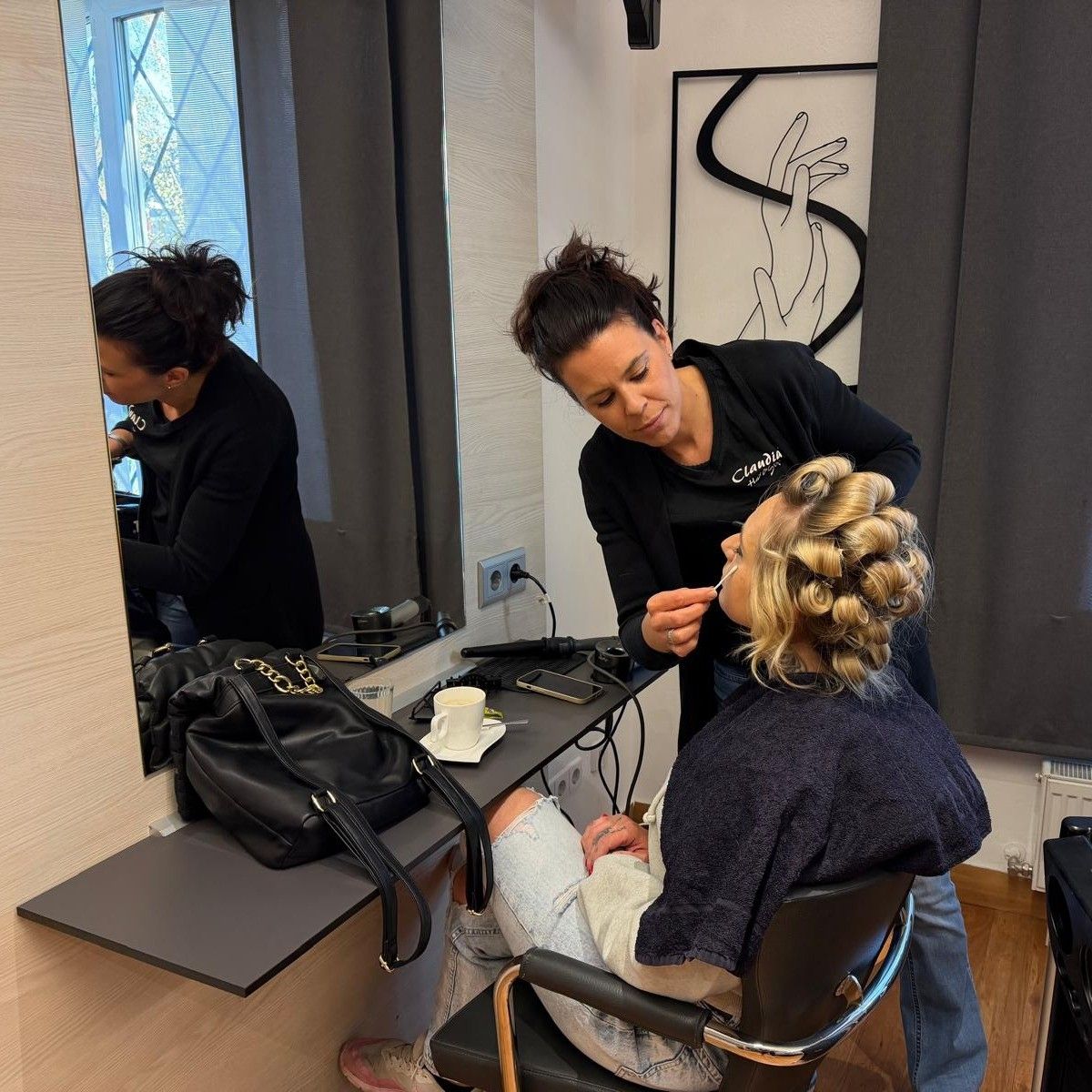 A woman is getting her makeup done by a professional in a salon. She sits in a chair while the professional stands behind her.