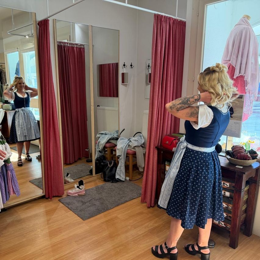 A woman is in a fitting room with mirrors, checking her outfit in the mirror. She has tattoos, a polka-dot skirt, and a blue blouse. Shoes and a bag are on the floor.
