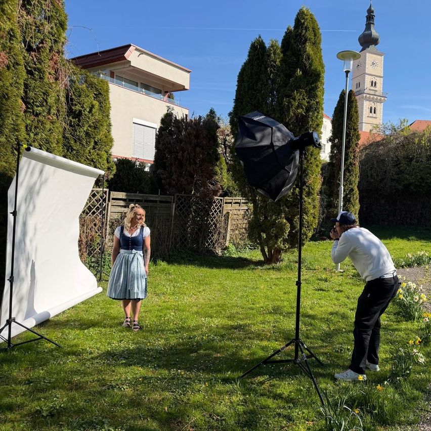 A woman in a traditional dress is posing for a photo in a garden with a photographer. The setup includes a backdrop, umbrella light, and a backdrop stand. Trees, a house, and a clock tower are in the background.