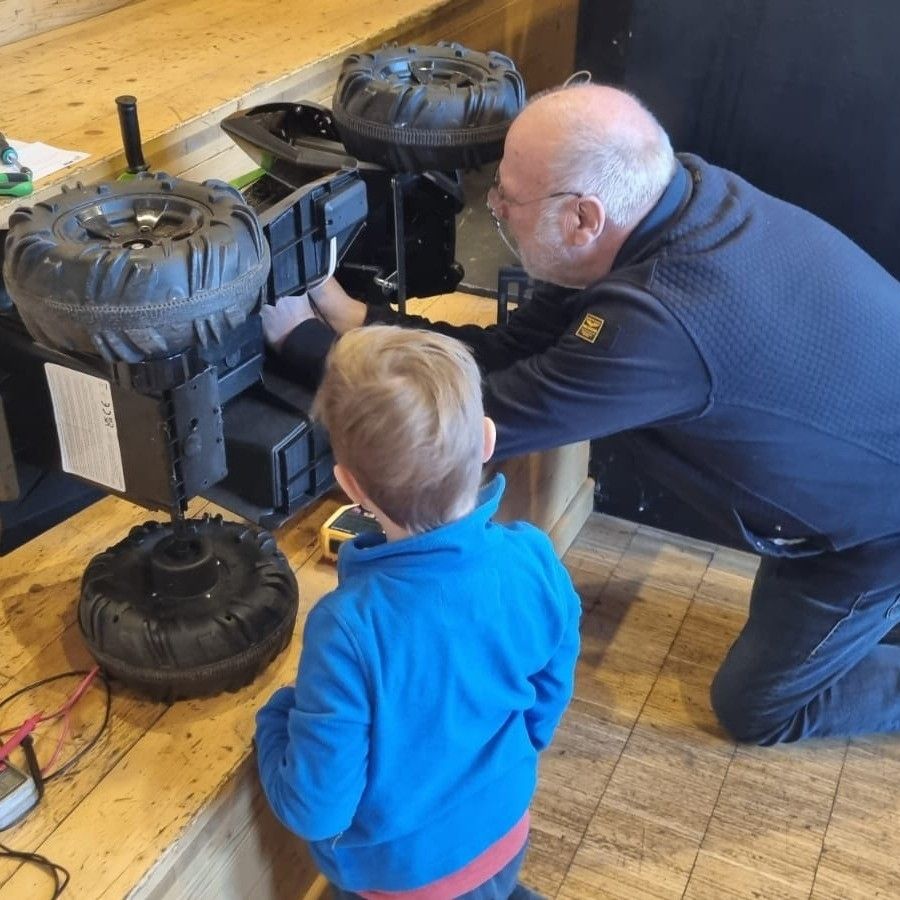 An older man helps a young boy inspect a vehicle with large tires. They are kneeling on a wooden floor.