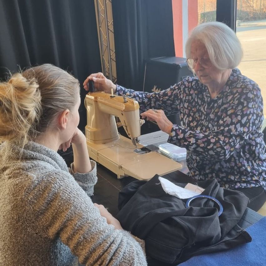 An older woman is teaching a younger girl how to use a sewing machine at a table. The girl is watching attentively.