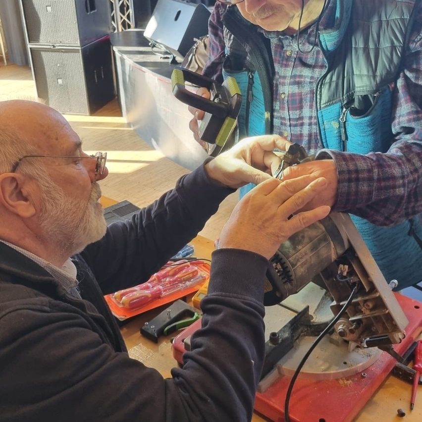 Two older men are working on a tool in a workshop. One man holds a tool in his hand while the other is fixing something on it.