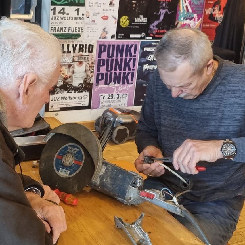 Two men work on an electrical project in a workshop, one holding a wire, the other a tool, with a poster reading Punk Punk Punk in the background.