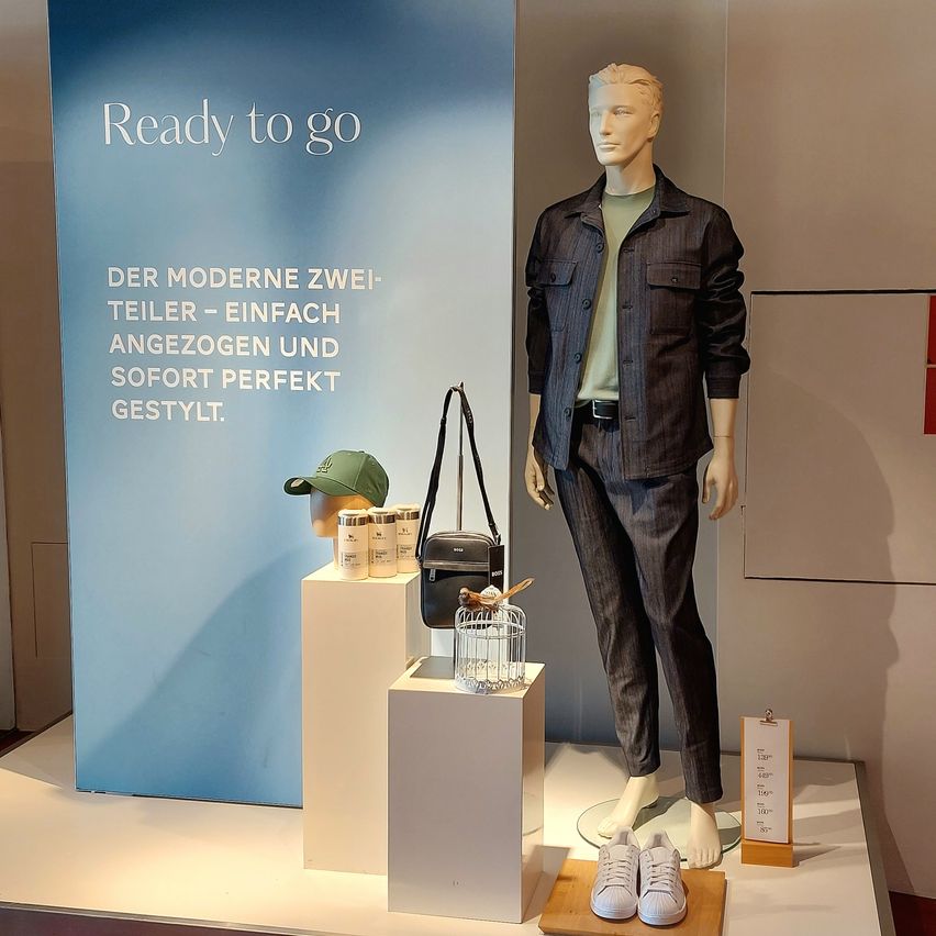 A mannequin displays a modern two-piece suit, with accessories on a platform. The sign reads 'Ready to go.'