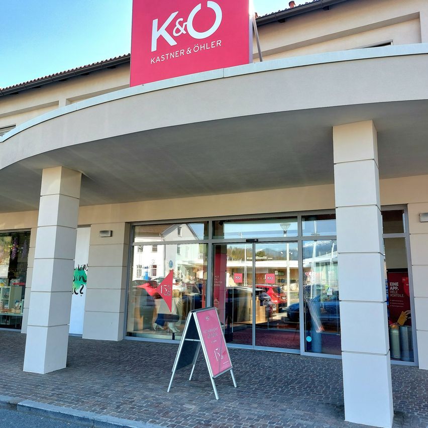 A K&O Kastner & Oehler store with a pink sign. The building has glass doors and windows. A signboard is in front.