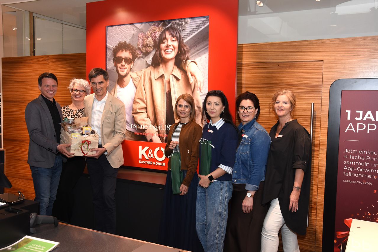 Five people pose for a photo in front of a large image, two holding gifts. Behind them, a man and woman are on a large photo. In front, a red banner with K&O and Kastner & Ohler.