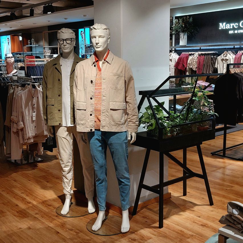 Two mannequins stand in a clothing store, one wearing a beige jacket and blue jeans, the other in a green jacket and white shirt. A plant display and clothes on hangers are in the background.