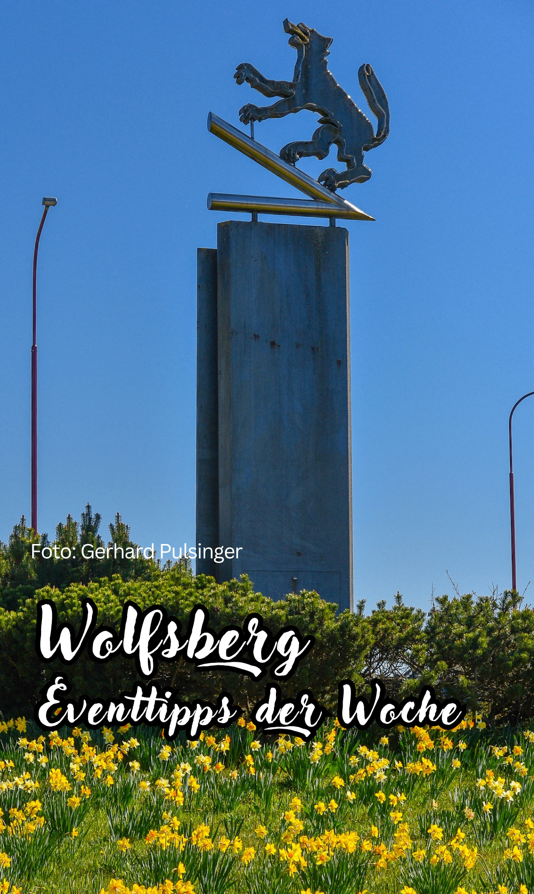Ein Betonmonument steht hoch, ein gelber Pfeil obenauf. Bäume umgeben es, mit blauem Himmel und Straßenlaterne sichtbar. Das Foto wird Gerhard Pulsinger zugeschrieben.