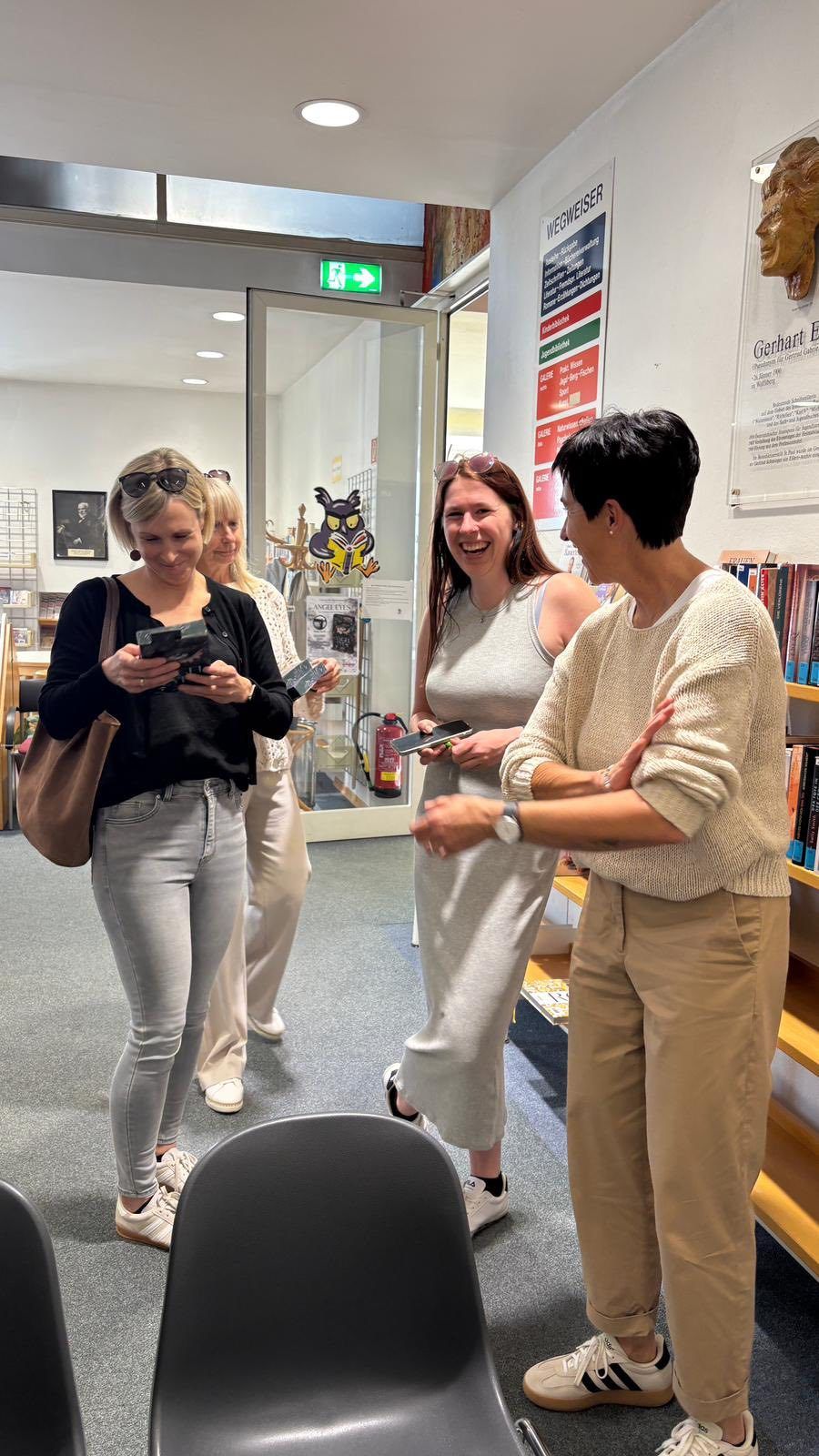 Vier Frauen stehen in einem Raum mit Büchern und einem Feuerlöscher. Zwei Frauen schauen auf ihre Telefone und eine lacht. Die andere Frau schaut zu.