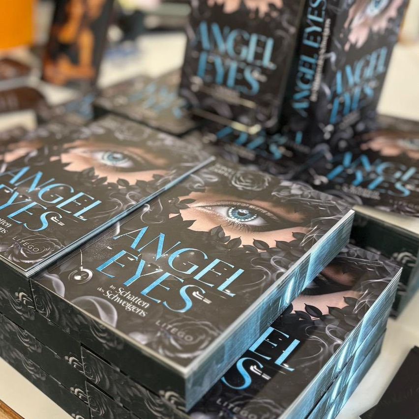 Stapel von 'Angel Eyes' Büchern sind auf einem Tisch ausgestellt. Die Bücher haben dunkle Cover mit blauen Augen und Blättern. 'Angel Eyes' steht in Weiß auf den Covern geschrieben.