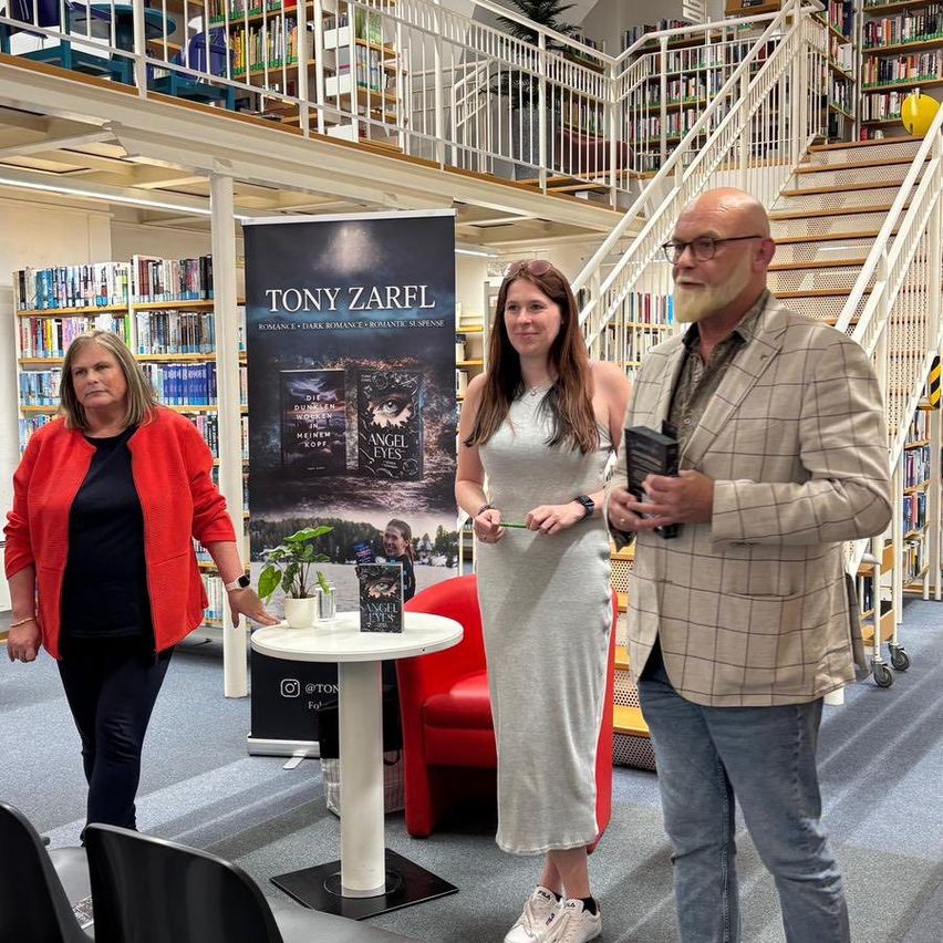 Eine Gruppe von Menschen steht in einer Bibliothek mit einem Banner hinter ihnen. Eine Frau schaut einen Mann an, der ein Buch hält.