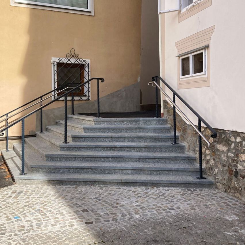 A building exterior with gray stone steps, black handrails, and a stone path. It has a window with a metal grill and a stone wall.