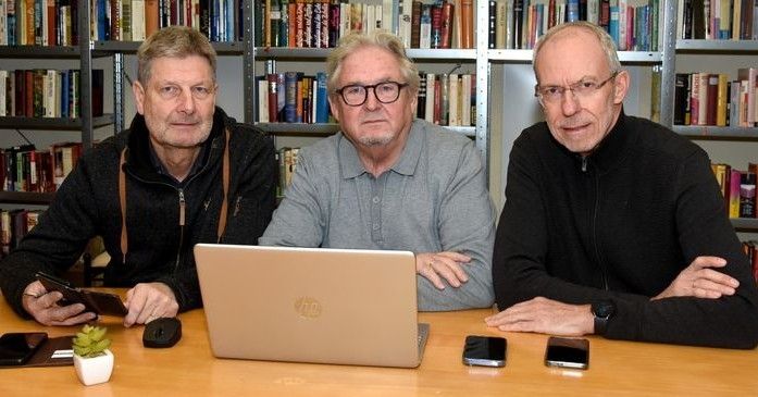Three men sit at a desk in a library. One holds a cell phone, another holds a laptop, and the third holds a wallet. They all have books in the background.
