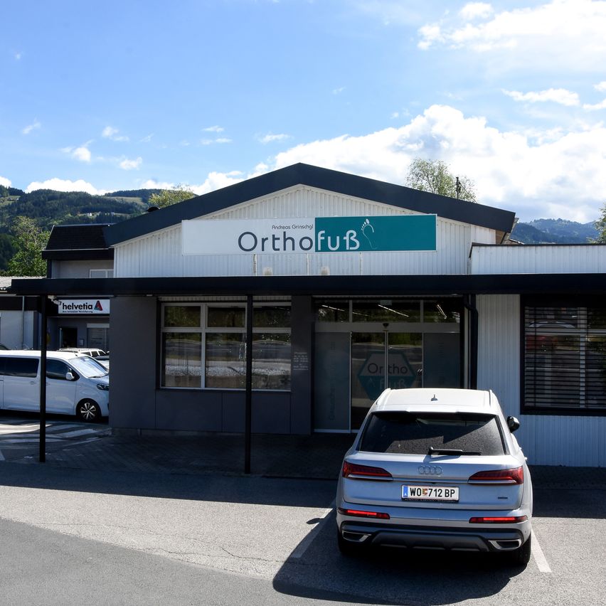 A silver Audi SUV is parked in front of the OrthofuB building with a white and green sign, set against a backdrop of mountains and a clear blue sky.