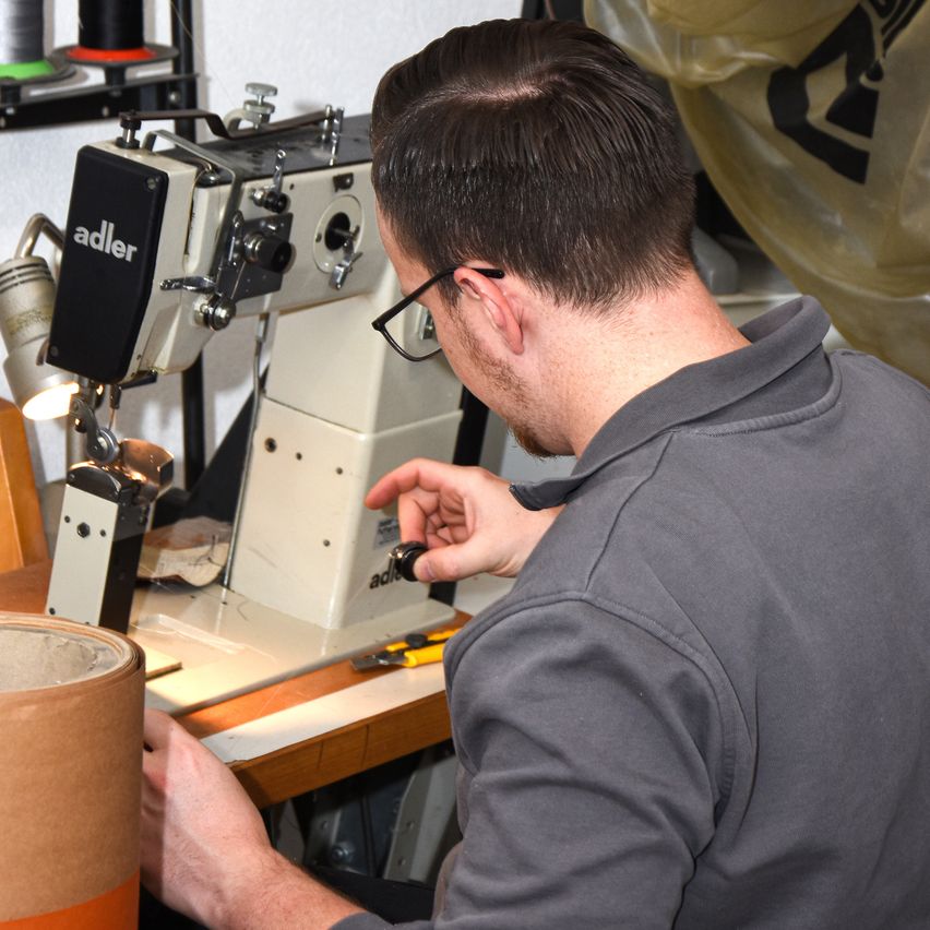 A man wearing glasses is working on an Adler sewing machine, holding a tool, and a rolled paper is on the desk.