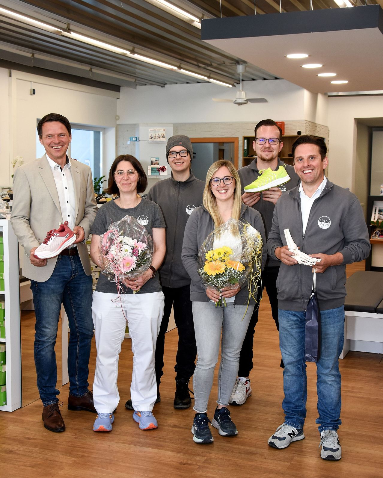 Six people stand in a room, smiling. The men hold shoes and bones. The women hold bouquets. One wears a ring. Wooden floors and white walls.