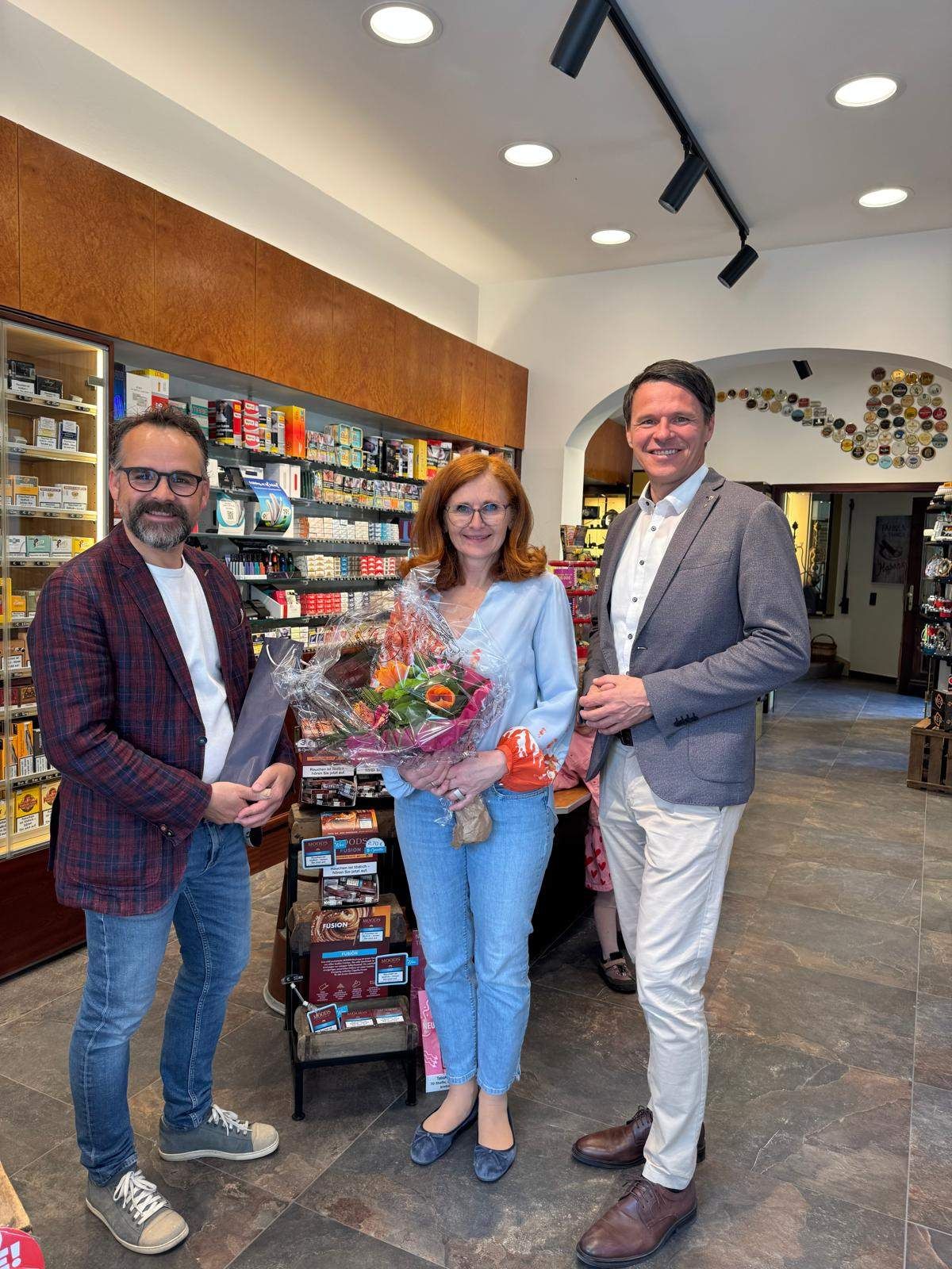 Three people stand in a shop, two men and a woman in the middle holding flowers. They appear to be posing for a photograph.